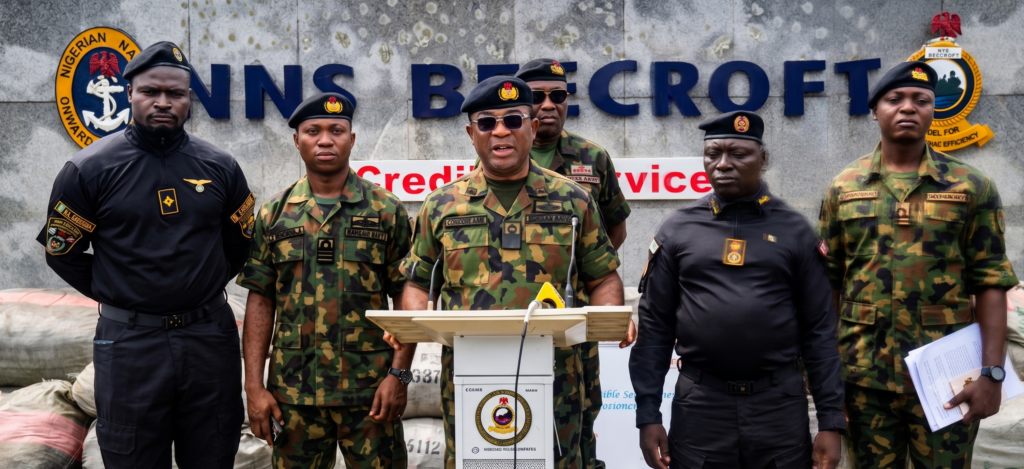 NNS BEECROFT Commander Commodore Aiwuyor Adams-Aliu speaks at a podium about the Falcon Eye surveillance system, flanked by naval and NDLEA officers next to piles of seized cannabis.